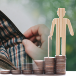 Eine Holzfigur eines Seniors mit Gehstock auf steigenden Münzstapeln neben einer geöffneten, leeren Geldbörse als Symbol für Rente und Altersarmut.