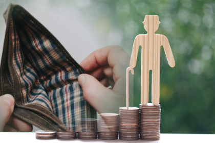 Eine Holzfigur eines Seniors mit Gehstock auf steigenden Münzstapeln neben einer geöffneten, leeren Geldbörse als Symbol für Rente und Altersarmut.